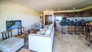 Una sala de estar con un sofá, una mesa de centro y un televisor. Hay una zona de cocina con taburetes de bar y un refrigerador.