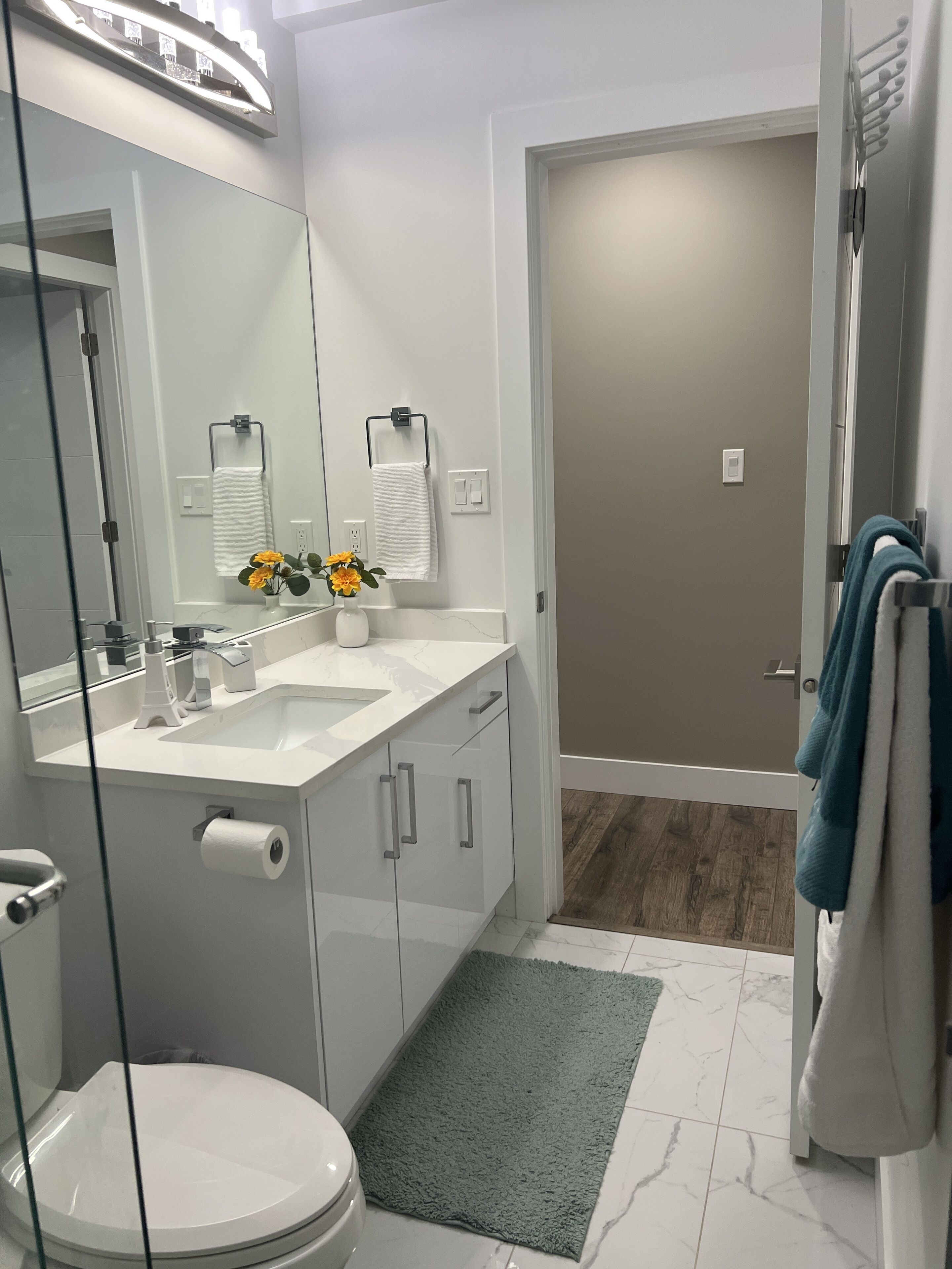 Un baño moderno con cabina de ducha de vidrio, un lavabo blanco y un inodoro. Hay una alfombra verde en el suelo y un perchero con una toalla colgada.