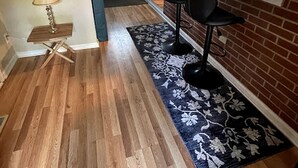 A kitchen with wooden flooring, a black rug, and a window with blinds.