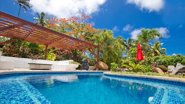Uma piscina com água azul cristalina, rodeada por plantas tropicais e uma pérgula de madeira.