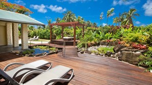 Um deck de madeira com espreguiçadeiras, uma banheira de hidromassagem e um jardim com plantas tropicais e um pequeno lago.