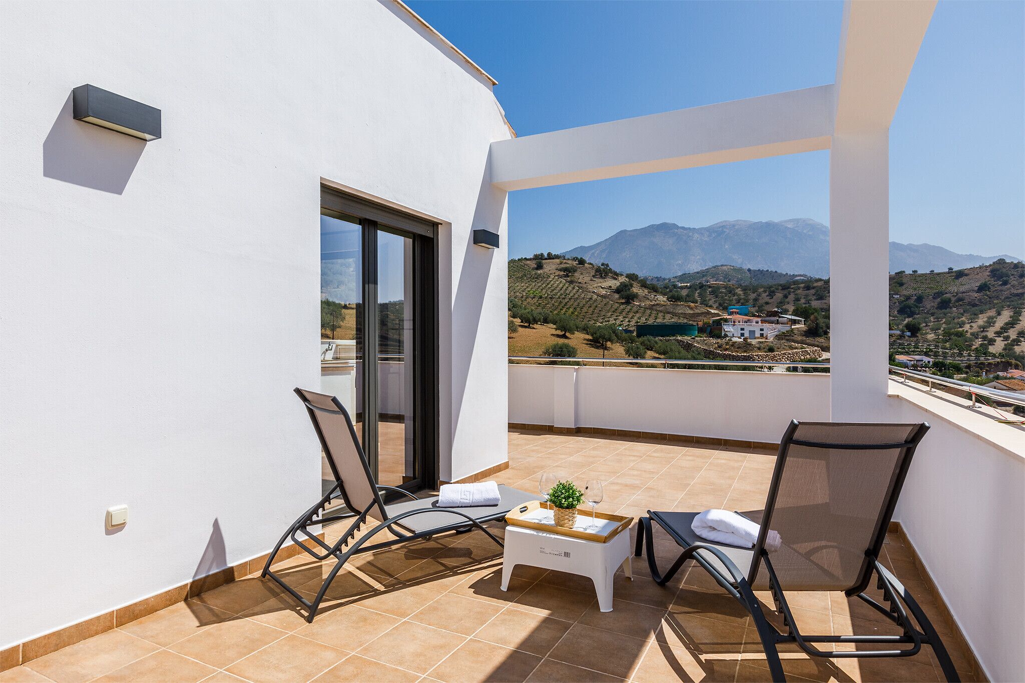 Un balcon avec deux fauteuils, une petite table ornée d’une plante et d’un verre, et une vue sur les montagnes et un village.