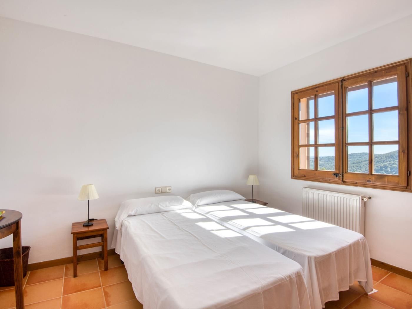 A room with two beds, a window, a radiator, and a small table with a lamp.