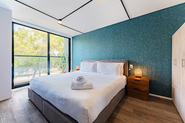 Un dormitorio moderno con una cama grande, vistas a árboles a través de un ventanal corredera y una pared azul con textura.