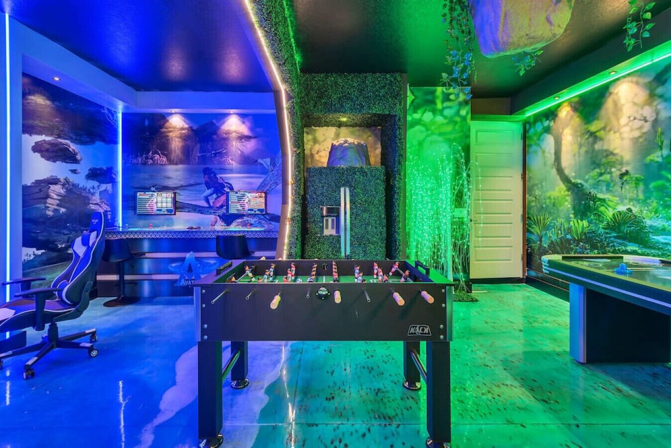 A room with a foosball table, a large screen displaying a game, and a seating area with a view of a mural.