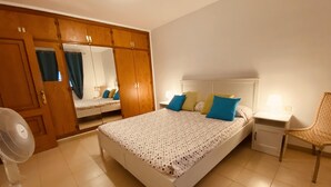 Une chambre à coucher avec un grand lit, une table de chevet, une chaise, un ventilateur et une armoire avec un miroir.