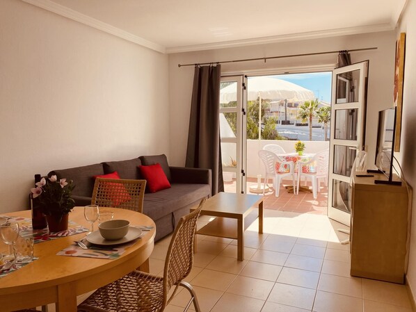 A living room with a sofa, a dining table with chairs, a TV stand, and a balcony with a table and chairs.