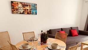 Un coin repas avec une table ronde en bois, six chaises en osier et un canapé gris agrémenté de coussins rouges. Un tableau représentant La Havane est accroché au mur.