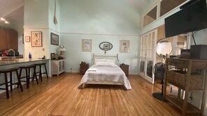 A bedroom with a wooden floor, a bed with a white headboard, a desk with a chair, and a TV mounted on the wall.