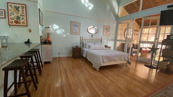 A bedroom with a wooden floor, a bed, a nightstand, a lamp, and a window with a view.