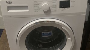 A front-loading white washing machine with a control panel and a label on the door. - 2 BR w/ AC APT near Grand Anse Beach -15 mins walk, 10 mins away from airport. (The Lime)