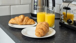 Una encimera de cocina con cruasanes en platos, una tetera y un exprimidor con vasos de zumo de naranja.