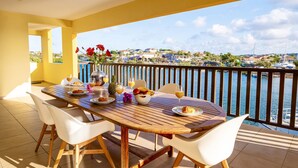 A wooden table set with breakfast items and a vase of red flowers on a balcony overlooking a marina with boats and houses.