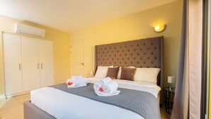 A hotel room with a large bed, a headboard, a wardrobe, an air conditioning unit, and a nightstand with a lamp.