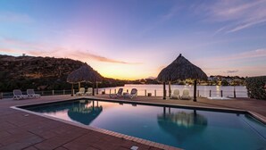 A swimming pool with lounge chairs and thatched umbrellas, surrounded by a landscaped area with a view of the water.
