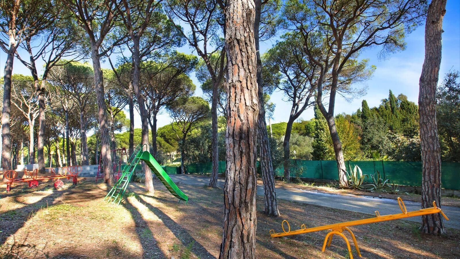 Un parque infantil con un tobogán verde y un sube y baja, en un parque con árboles altos.