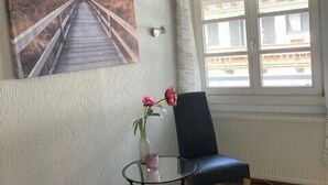 Una habitación con un cuadro de un puente, una silla, una mesita con un florero y una ventana con cortinas.