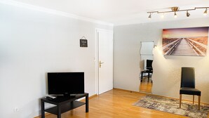 Un salón con una televisión de pantalla plana sobre un soporte negro, suelo de madera, una silla negra y un cuadro de una playa en la pared.