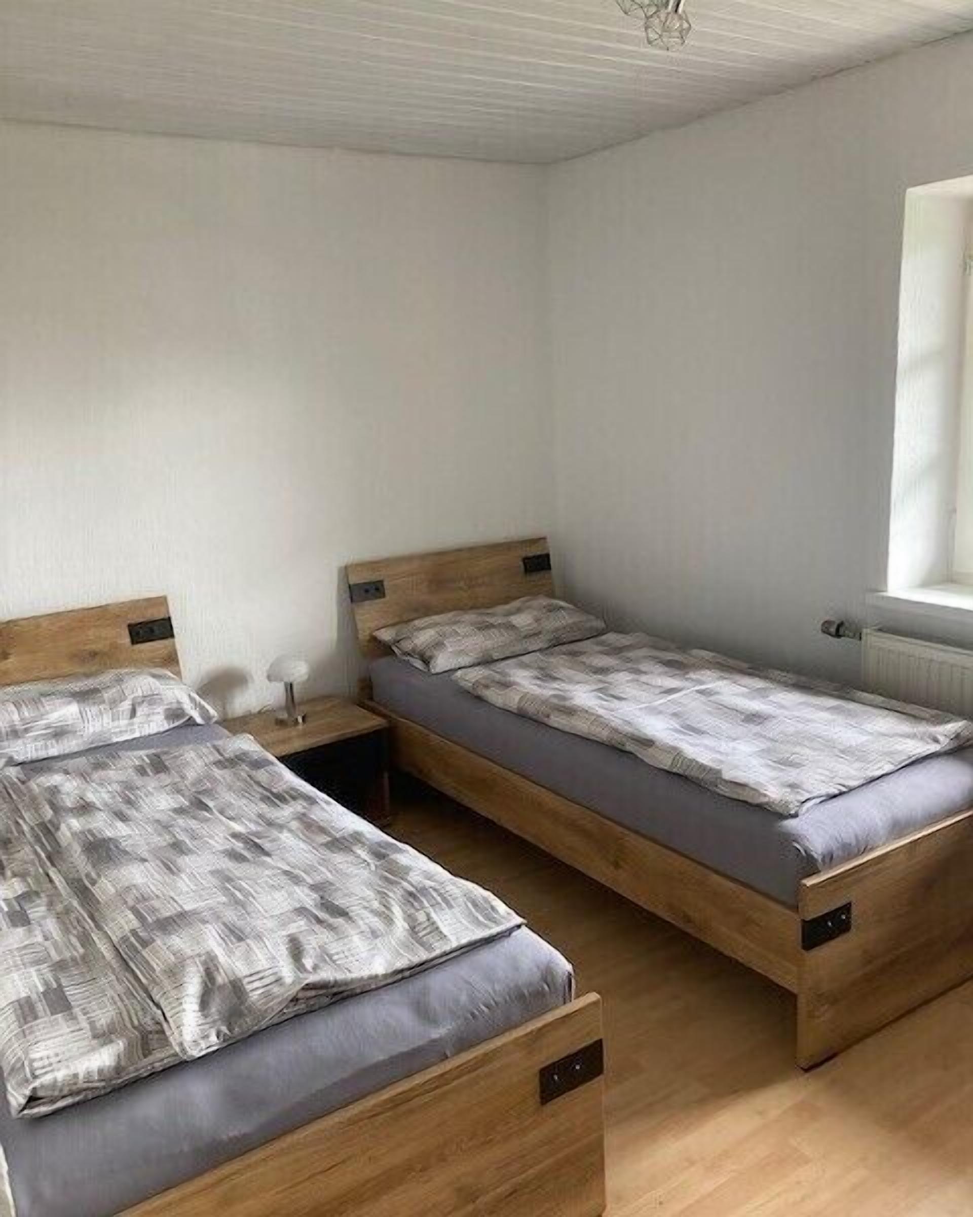 A room with two wooden beds, each with a grey mattress and matching bedding. There is a small bedside table with a lamp on one side. The room has white walls and a window on the right side.
