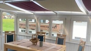 Un invernadero con una mesa y sillas de madera, ventanas amplias y vistas de edificios.