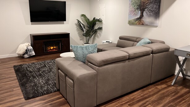 A modern living room with a sectional sofa, a fireplace, a flat-screen TV, and a painting on the wall. - Feel at home in This 2 Bedroom Basement Suite (Kamloops)