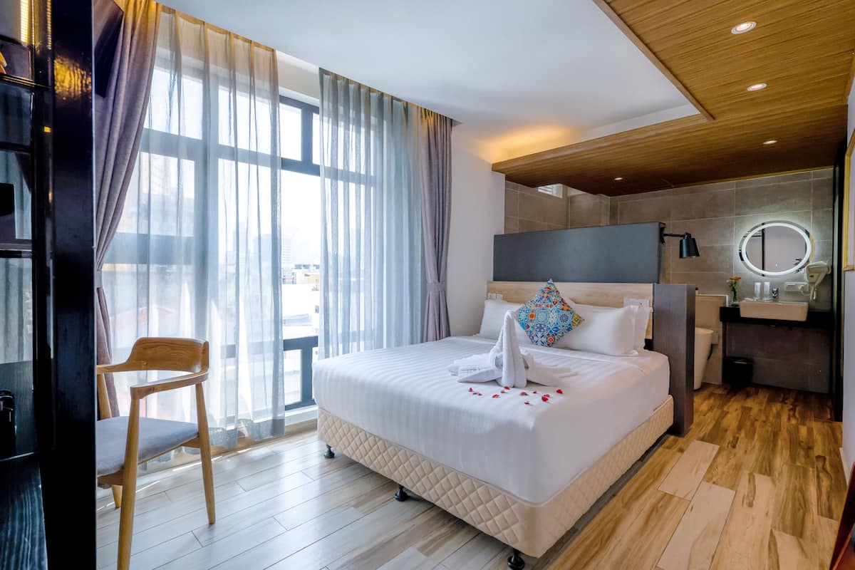 A hotel room with a large bed, a wooden chair, a bathroom with a mirror, and a view through the window.