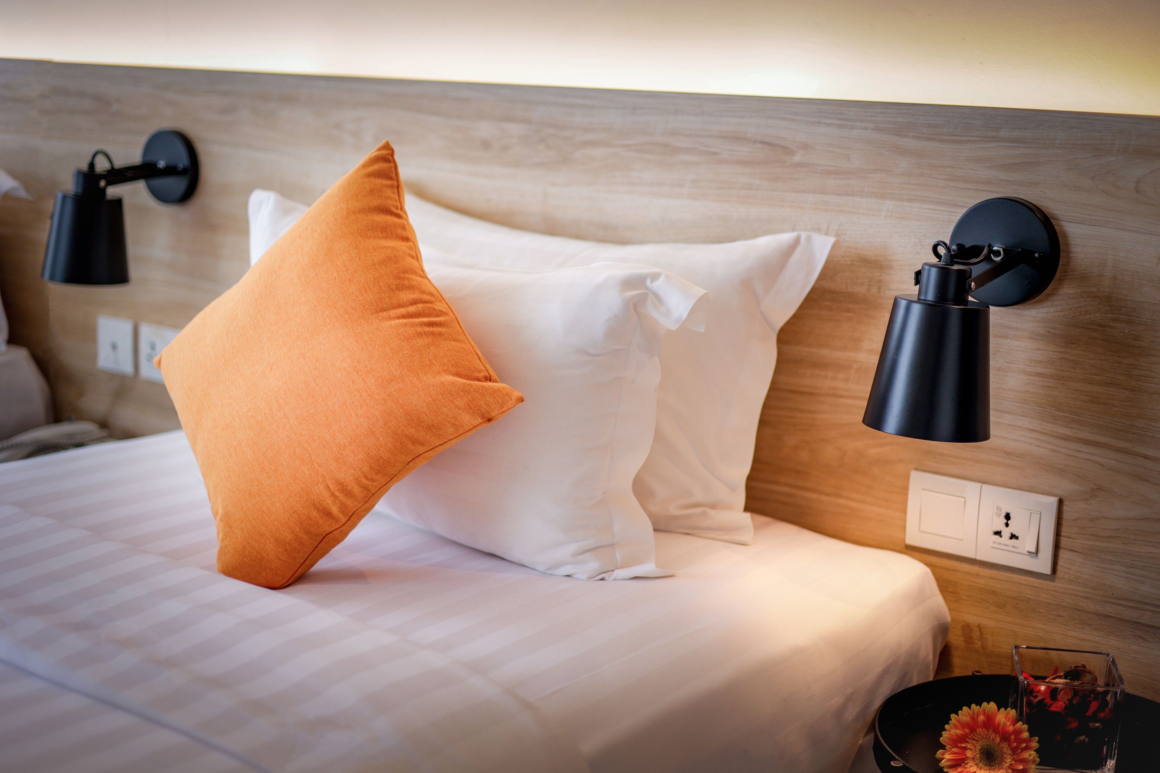 Una cama bien hecha con almohadas blancas y naranjas, una mesita de noche con un vaso y flores, y lámparas fijadas a la pared.