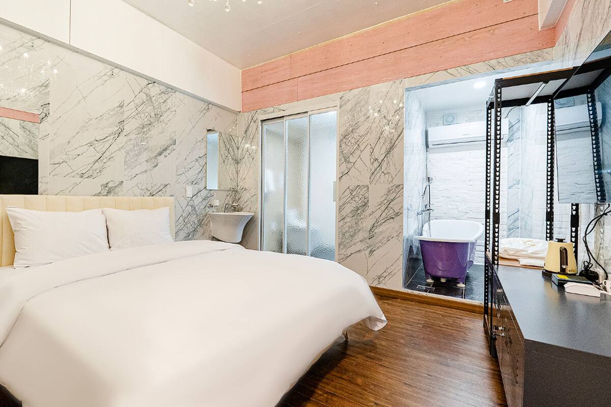 Quarto de hotel moderno com parede de mármore, uma cama grande, banheiro com banheira roxa e uma escrivaninha com computador.