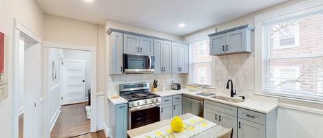 A compact kitchen with grey cabinets, a microwave, stove, sink, and a window with blinds.