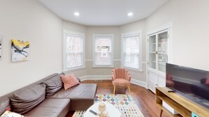 Un salon avec un canapé en cuir marron, un fauteuil rose, un téléviseur posé sur un meuble en bois et un tapis à motifs.