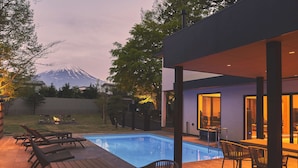 Zona de piscina moderna al aire libre con sillones de descanso y una terraza de madera, rodeada por una casa con vistas a la montaña.