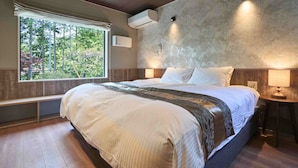 Un dormitorio con una cama grande, una banca, un ventanal con vistas a árboles, un equipo de aire acondicionado y una lámpara fijada a la pared.