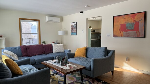 A modern living room with a blue sofa set, a wooden coffee table, and a colorful rug. There is a kitchen visible in the background, and a large abstract painting on the wall. - Lovely cottage with fenced in back yard (Wausau)