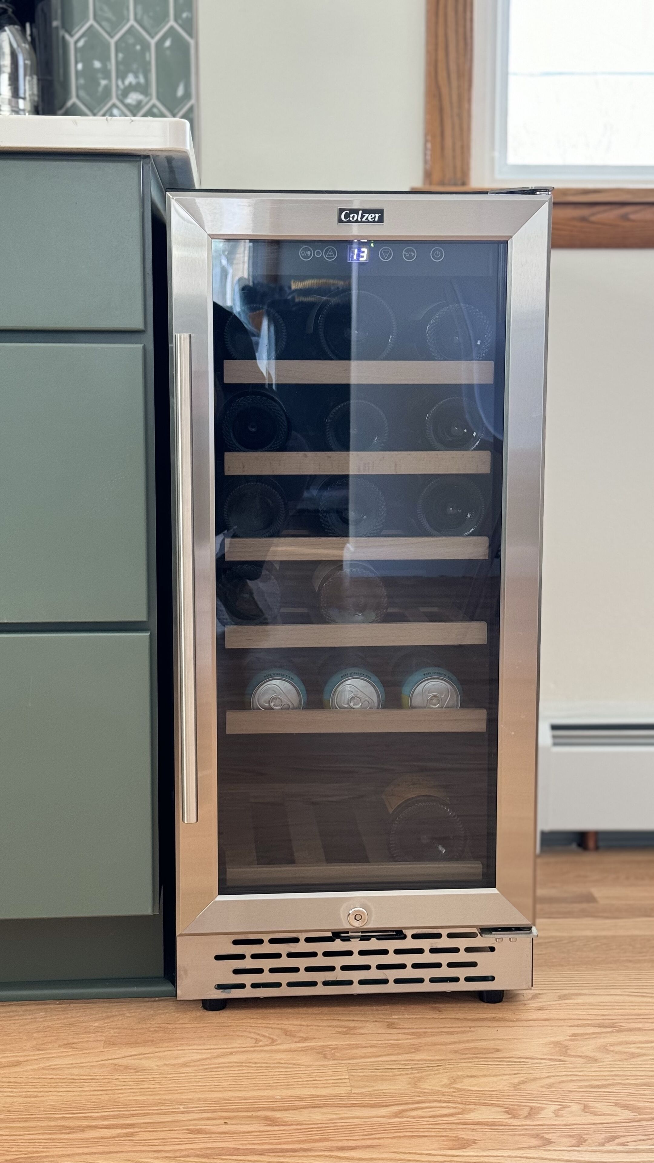 A Colzer wine cooler with a glass front and multiple shelves, placed in a kitchen with green cabinets and a wooden floor.