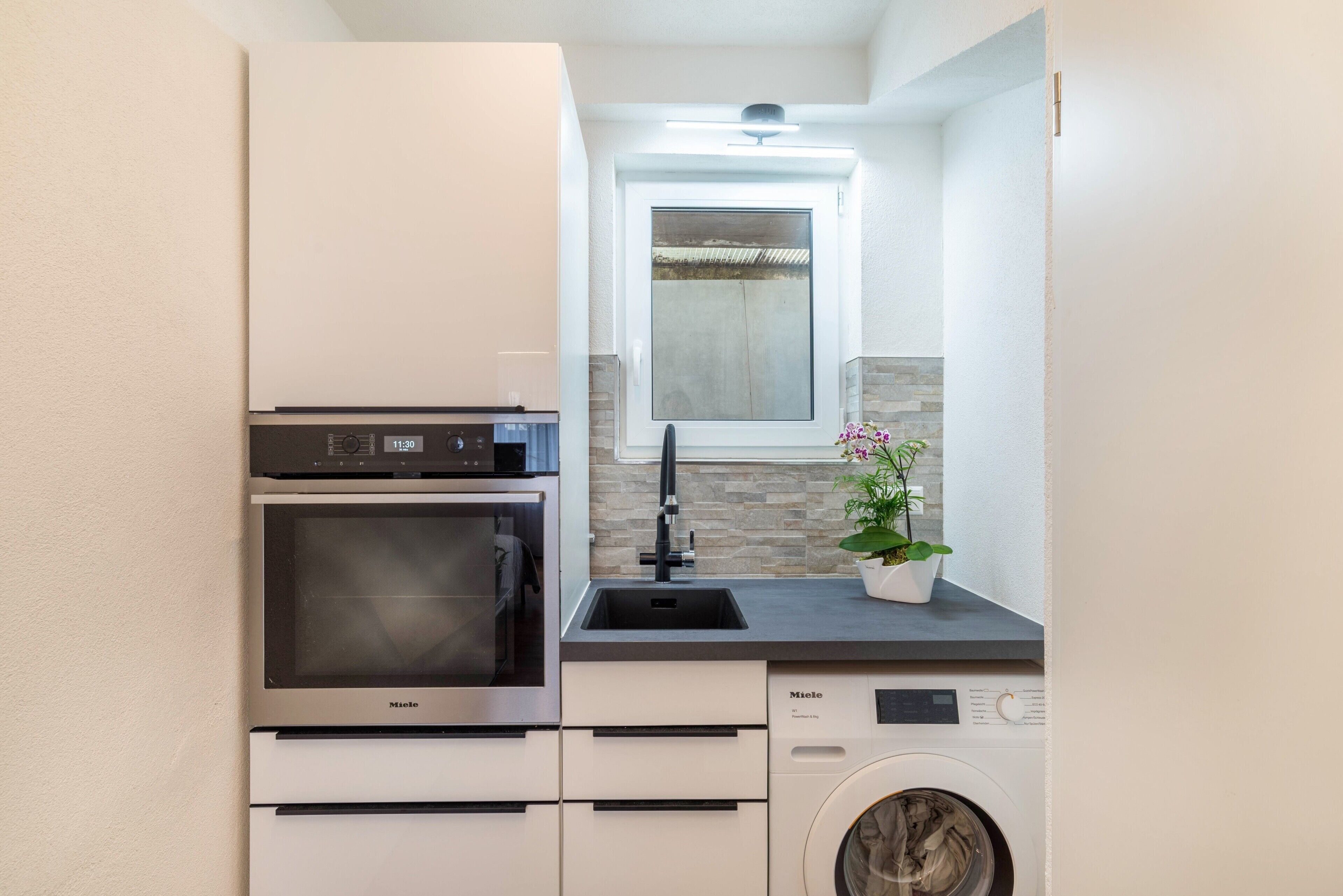 A compact kitchen with a built-in oven, a washing machine, and a window above the sink.