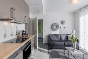 A modern kitchen with a built-in oven, a dining area with a black sofa and a coffee table, and a living room with a grey sofa and decorative wall art.