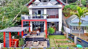 Un edificio de dos pisos con balcón, un espacio al aire libre cubierto y un jardín con bancales elevados.