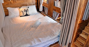 Una cama bien hecha con sábanas blancas, un cojín con forma de gato de dibujos animados, cabecera de madera y cortinas.