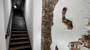 Una escalera con escalones de madera que ascienden, flanqueada por paredes blancas y un muro de ladrillo parcialmente visible.