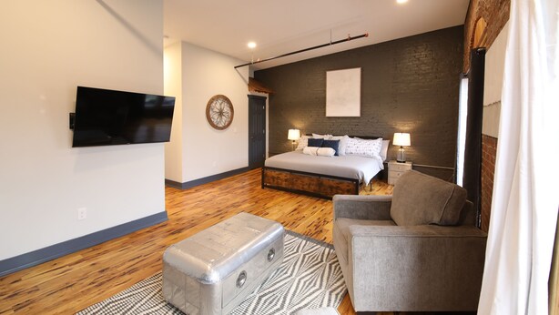 Un dormitorio moderno con cama, televisión, piso de madera y un sillón gris.