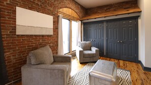 Una habitación con pared de ladrillo, un ventanal con cortinas, un sillón gris, un sillón más pequeño, un banco de madera y un armario gris.