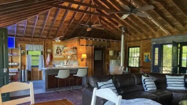 A cozy cabin interior with a dining area, a living room with a sofa and chairs, a kitchen area, and a large window providing natural light. - Airstream Glamping in The Adirondack Foothills (Argyle)