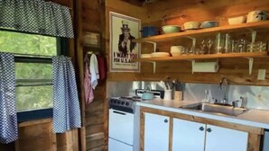 A compact kitchen with wooden cabinets, a stove, and a sink. There are shelves above the counter holding dishes and glasses. - Airstream Glamping in The Adirondack Foothills (Argyle)