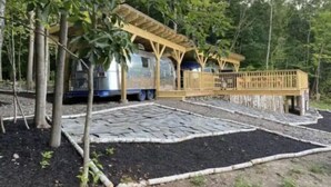 A covered outdoor area with a wooden deck, stone flooring, and a metal structure in the background. - Airstream Glamping in The Adirondack Foothills (Argyle)