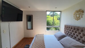 Une chambre à coucher comprenant un lit, un téléviseur fixé au mur, une fenêtre donnant sur la verdure et un élément décoratif mural.