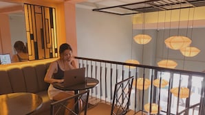 Een vrouw gebruikt een laptop aan een ronde tafel in een kamer met een balkon, een patroon vloer en hangende lampen.