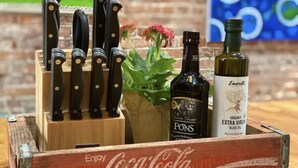 Une boîte en bois ornée du logo Coca-Cola, un ensemble de couteaux, une bouteille de vin Pons et une bouteille d’huile d’olive Emirelli.