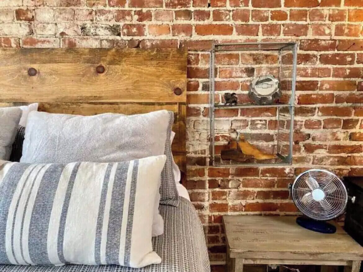 A bed with striped pillows, a wooden headboard, a brick wall, a small shelf with decorative items, and a fan on a wooden table.