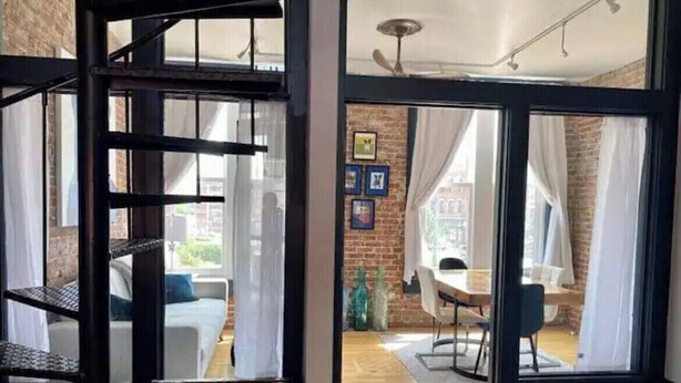 Un escalier mène à une pièce dotée d’un plancher en bois, d’un coin repas avec une table et des chaises, et offrant une vue sur l’extérieur grâce à de grandes fenêtres.
