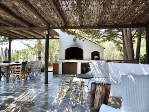 An outdoor patio with a stone floor, wooden chairs, a table, a pergola with a thatched roof, and a stone oven.